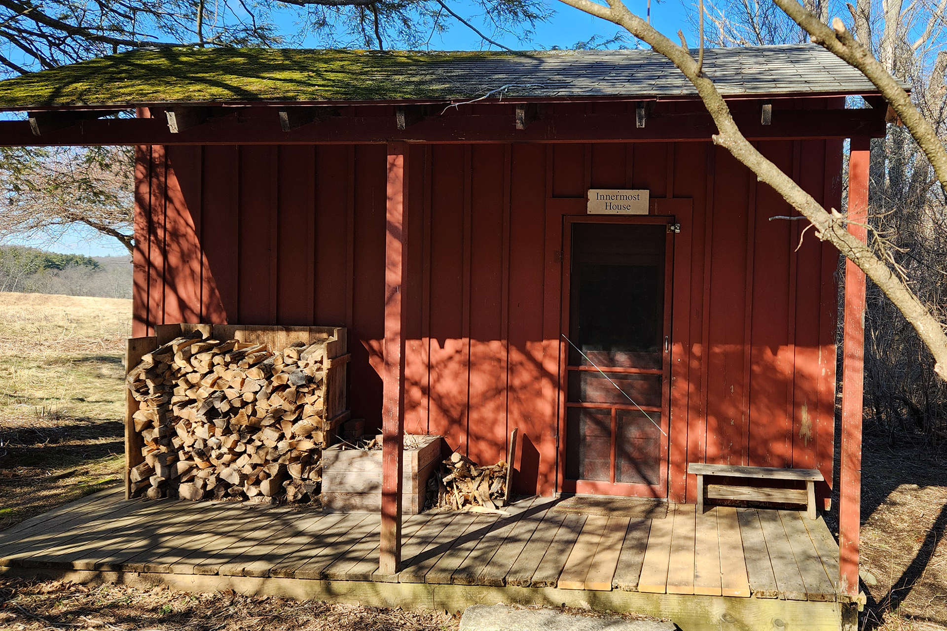 Picture of a brick red cabin with firewood stacked outside to the left of the door