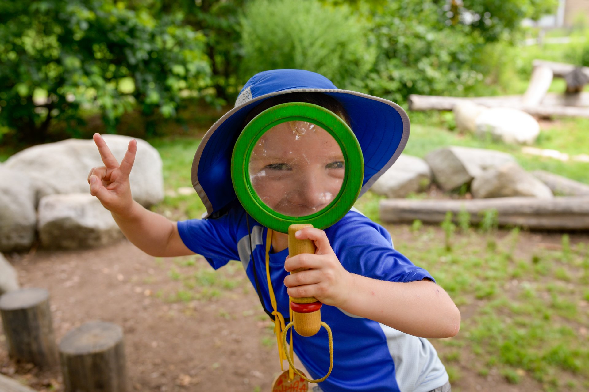 A camper wearing a blue shirt and sunhat, making the peace sign with one hand and holding an oversized magnifying glass in front of his face with the other