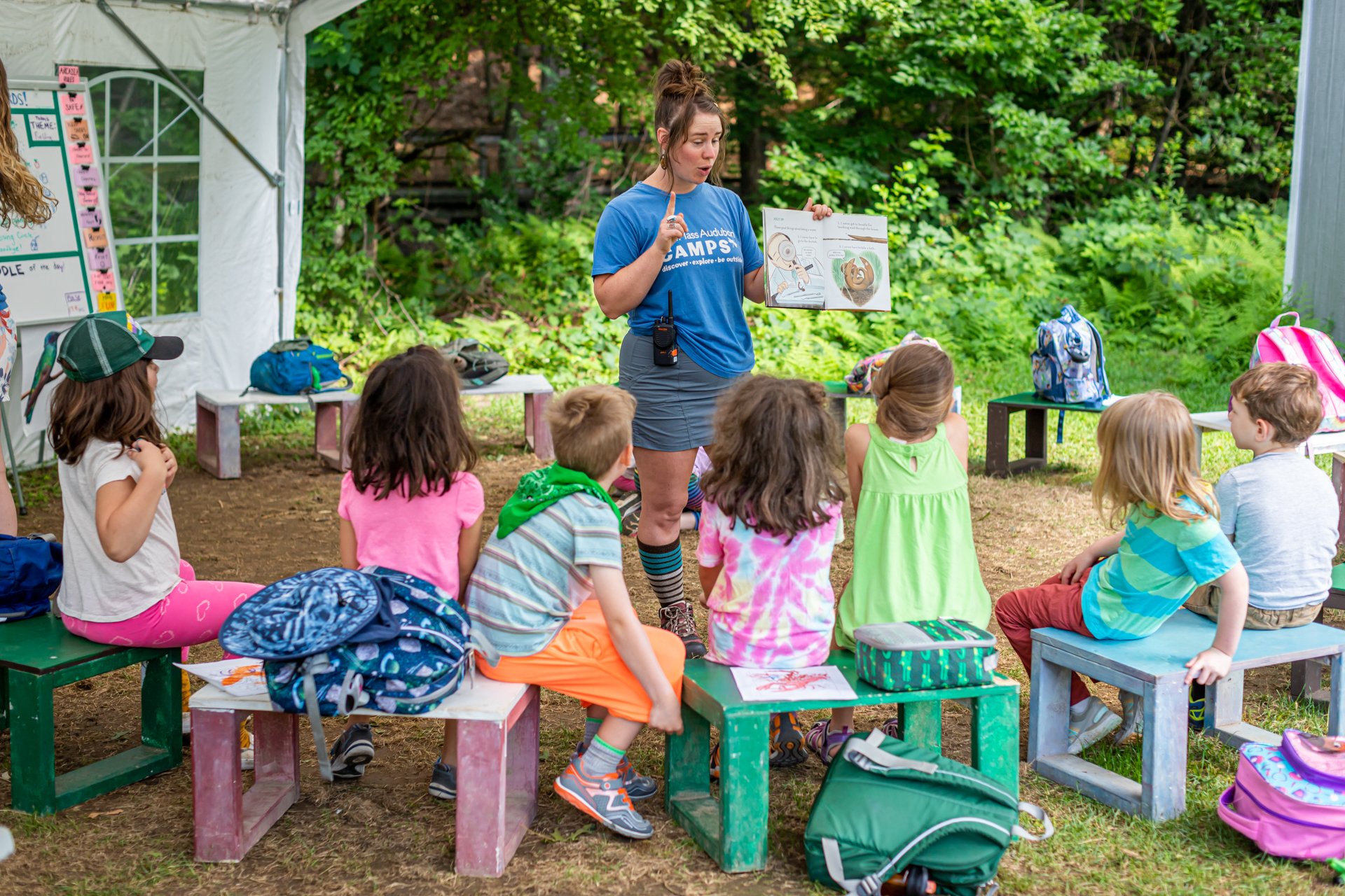 A group of campers at Arcadia Nature Camp seated in a circle, listening attentively to a counselor reading from a picture book with a radio clipped to her waistband