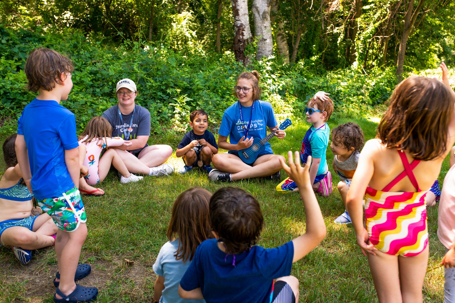 Drumlin Farm campers seated in the grass in a singing circle; a counselor plays a ukelele