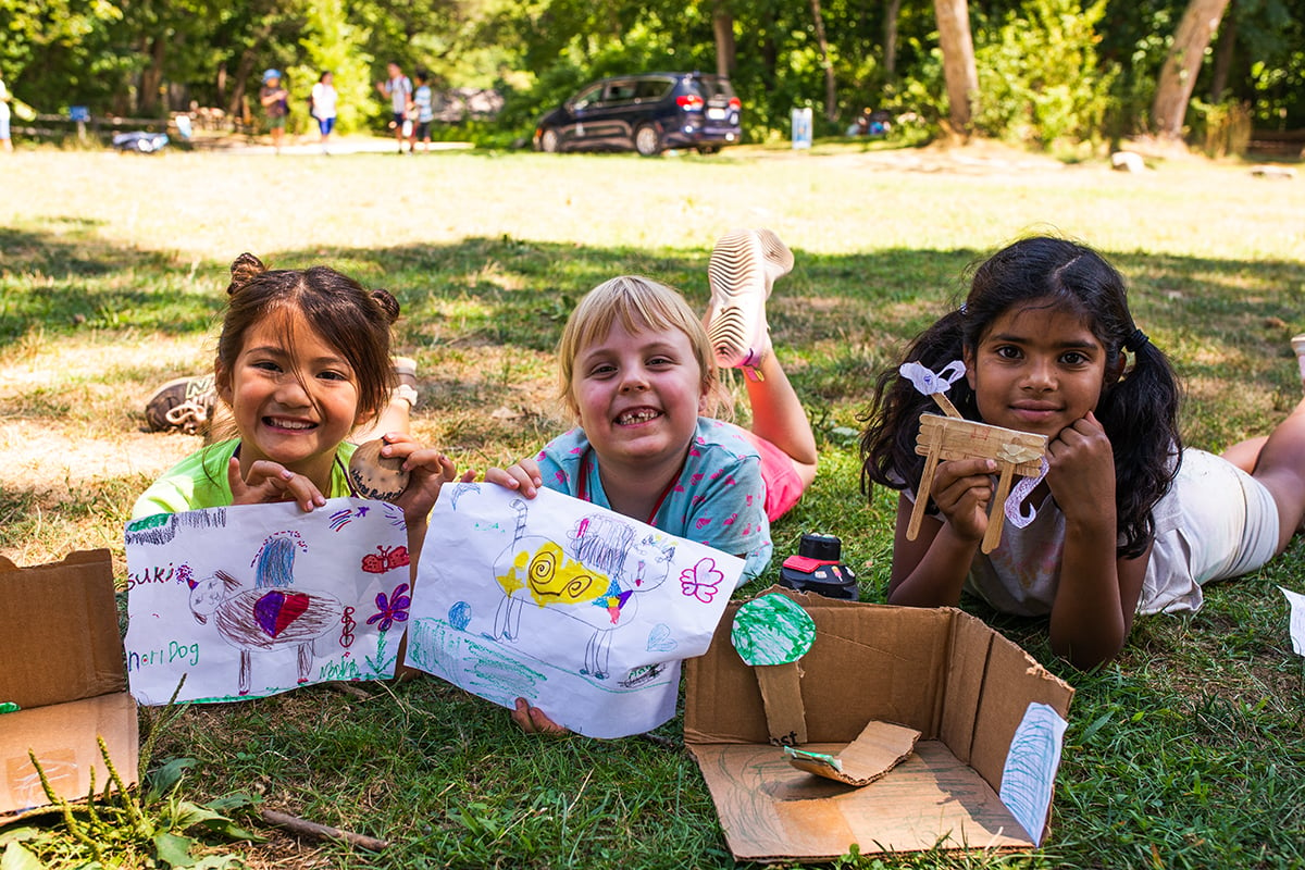 3 young campers laying on the ground showing of drawings and creations