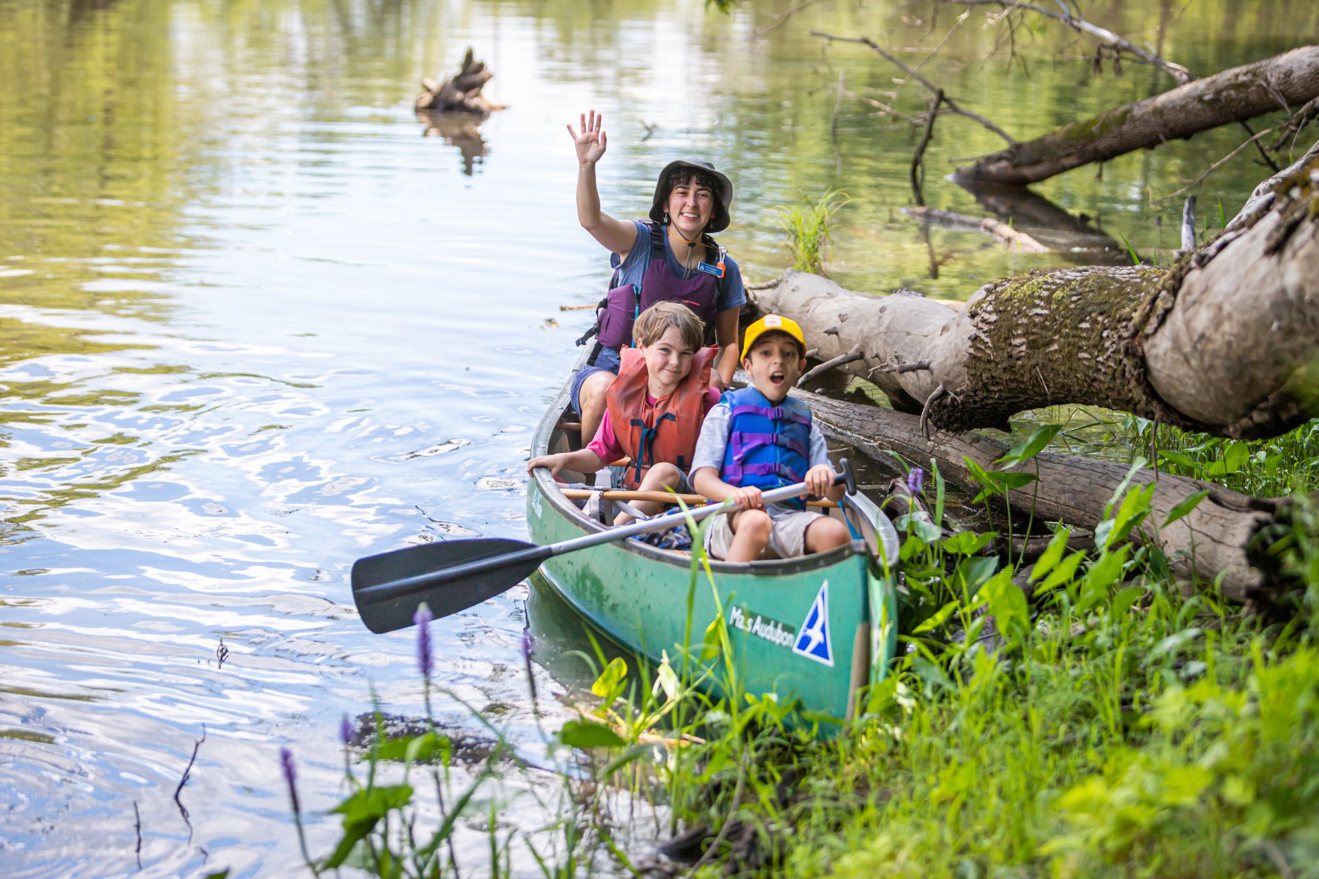 Two Arcadia campers and a counselor, wearing PFD life vests, wave from a canoe at the edge of a river