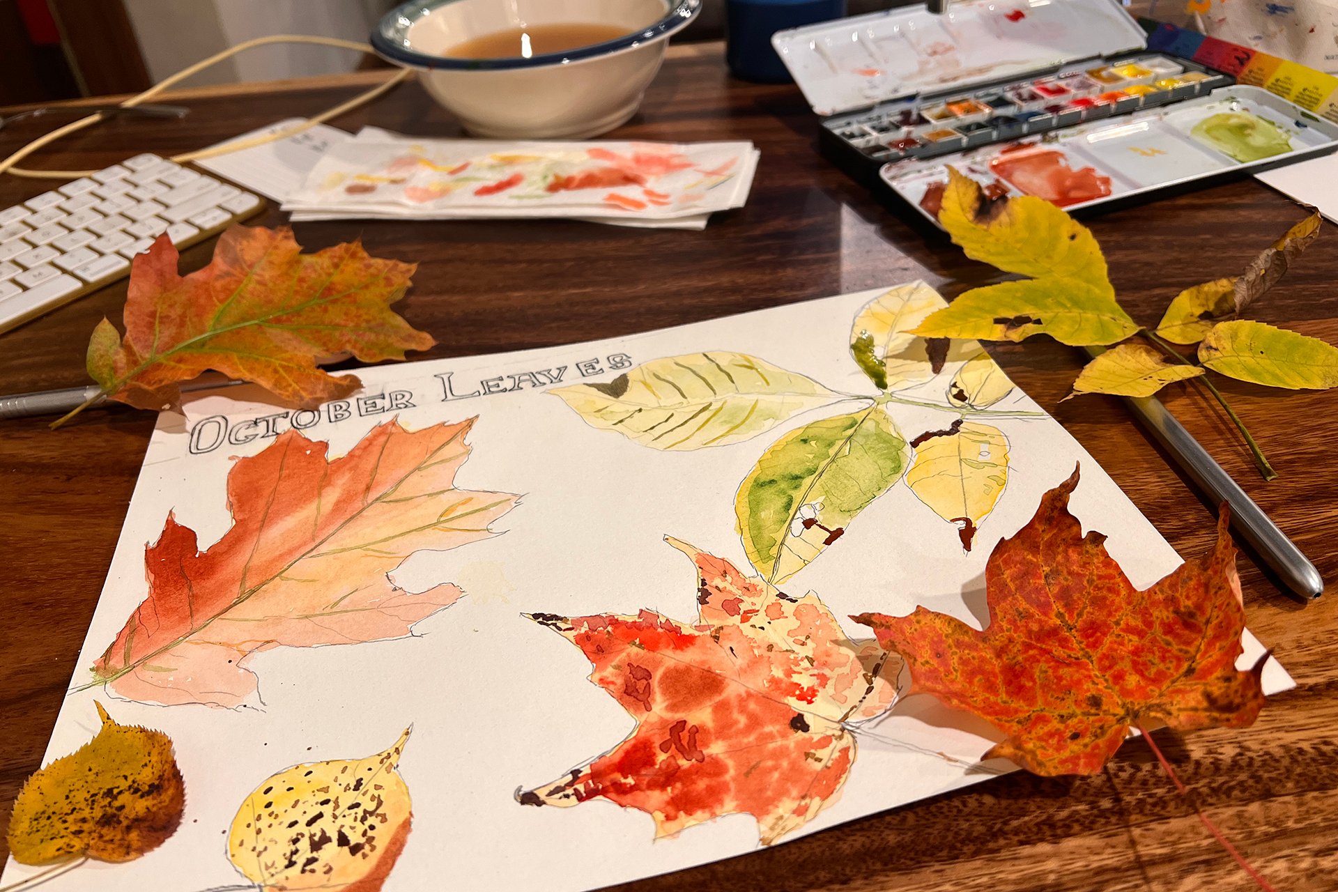 Piece of paper with watercolor of fall leaves on a table with art supplies