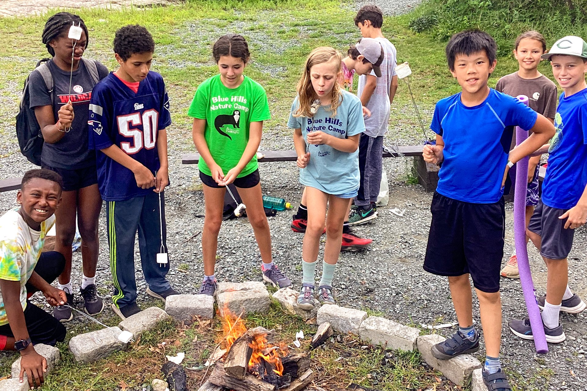 A group of Blue Hills campers standing around an open campfire toasting marshmallows with long, metal roasting forks